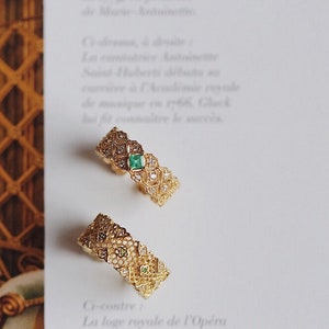 May include: Two gold rings with intricate filigree designs. One ring features a green gemstone in the center, while the other ring has a smaller green gemstone. Both rings are set against a white background.
