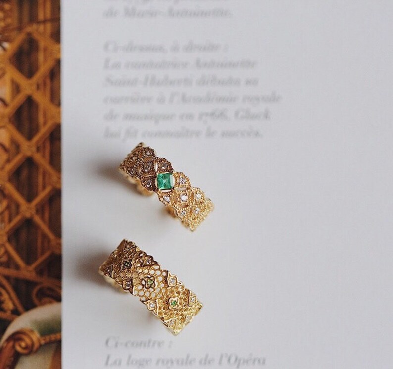May include: Two gold rings with intricate filigree designs. One ring features a green gemstone in the center, while the other ring has a smaller green gemstone. Both rings are set against a white background.
