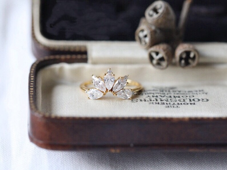 Puede incluir: Un anillo de oro con un grupo de piedras claras en forma de marquesa engastadas en una banda delicada y curva. El anillo se muestra en una caja de anillo de terciopelo marr&oacute;n.