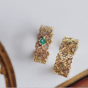 May include: Two gold rings with intricate filigree designs. One ring features a square emerald gemstone, while the other ring has smaller green gemstones.