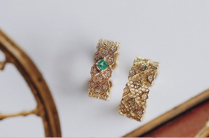 May include: Two gold rings with intricate filigree designs. One ring features a square emerald gemstone, while the other ring has smaller green gemstones.