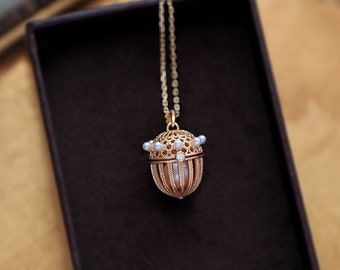 Pode incluir: Um colar de ouro com um delicado pingente de gaiola adornado com pequenas pérolas brancas. O pingente tem a forma de uma bolota e apresenta um design delicado e intrincado.