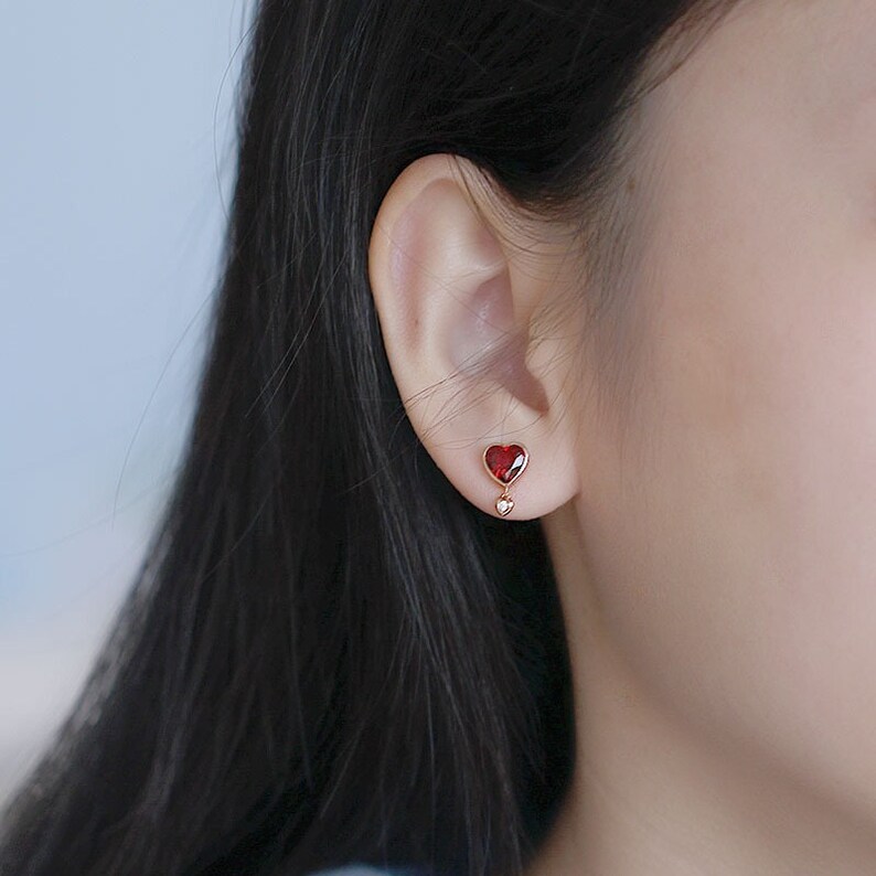 May include: A pair of gold heart-shaped earrings with a red gemstone in the center of the larger heart and a smaller clear gemstone in the center of the smaller heart.