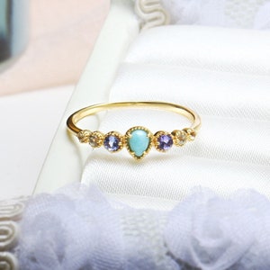 May include: A gold ring with a turquoise teardrop-shaped stone in the center, surrounded by smaller stones in shades of purple and white.