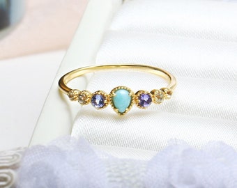Può includere: Un anello in oro con una pietra turchese centrale a forma di pera, affiancata da due pietre rotonde viola e due pietre chiare più piccole. Le pietre sono incastonate in una sottile fascia dorata con una texture metallica perlata.