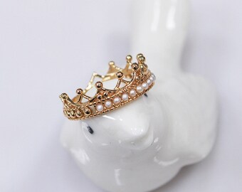Può includere: Un anello a forma di corona dorata con piccole perle bianche intorno alla base.