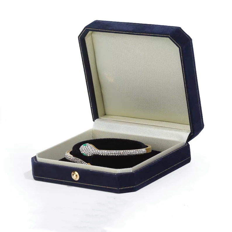 Op de afbeelding: Een blauwe fluwelen juwelendoos met een gouden sluiting, open om een wit satijnen interieur te onthullen. Een diamanten en smaragden slangenarmband wordt in de doos getoond.