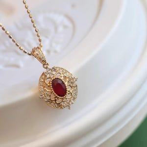 May include: A gold-coloured pendant necklace with an oval, deep red gemstone. The pendant has a detailed filigree design and small, clear gemstones. The delicate chain complements the ornate style of the pendant. A classic piece of jewellery.