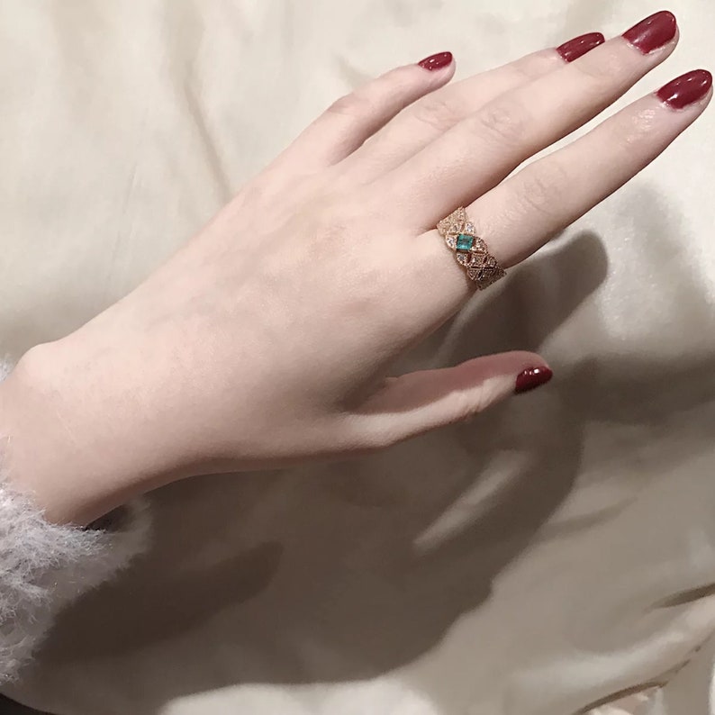 May include: A gold ring with a delicate design featuring a green gemstone. The ring is worn on the middle finger of a hand with red nail polish.