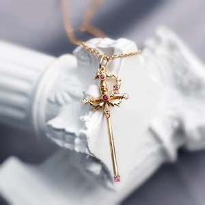May include: A gold necklace with a pendant shaped like a sword with a scorpion design. The sword is adorned with red and white gemstones.