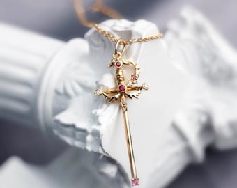 May include: A gold pendant necklace shaped like a sword, featuring a long, slender blade, a crossguard with ornate details, and a hilt. The piece is set with small, round, red gemstones and clear, sparkling white stones throughout.