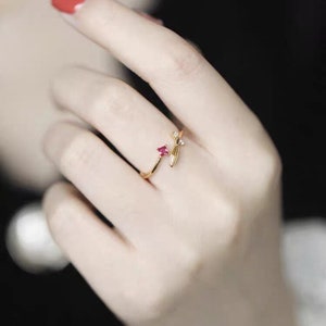 Puede incluir: Un anillo de oro con una piedra preciosa roja y un delicado dise&ntilde;o de hoja. El anillo se lleva en un dedo.