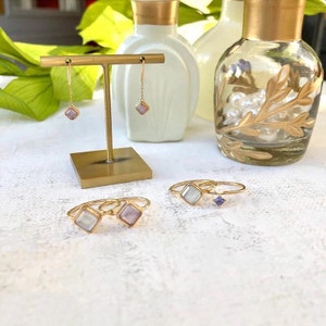 May include: A set of gold jewelry featuring a pair of dangle earrings with square pink stones and three rings with square stones. Two of the rings have white stones and one ring has a purple stone.