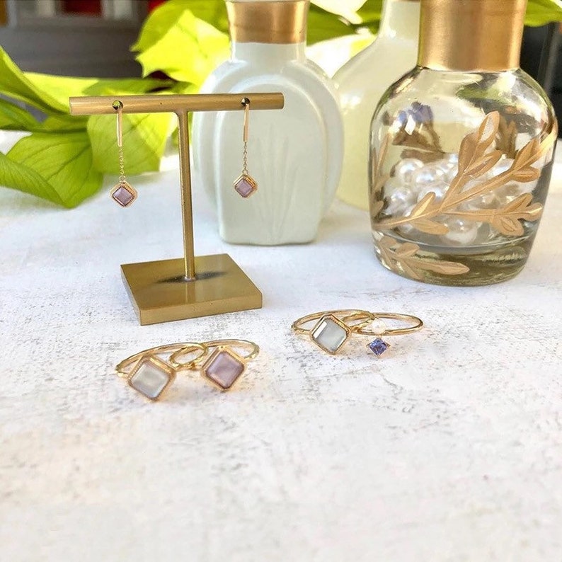 May include: A set of gold jewelry featuring a pair of dangle earrings with square pink stones and three rings with square stones. Two of the rings have white stones and one ring has a purple stone.
