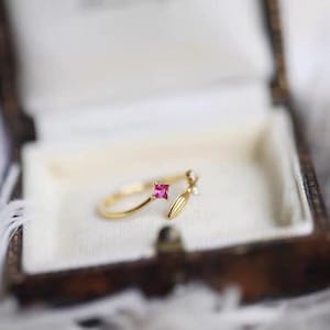 May include: A gold ring with a pink square gemstone and a delicate leaf design. The ring is open and adjustable.