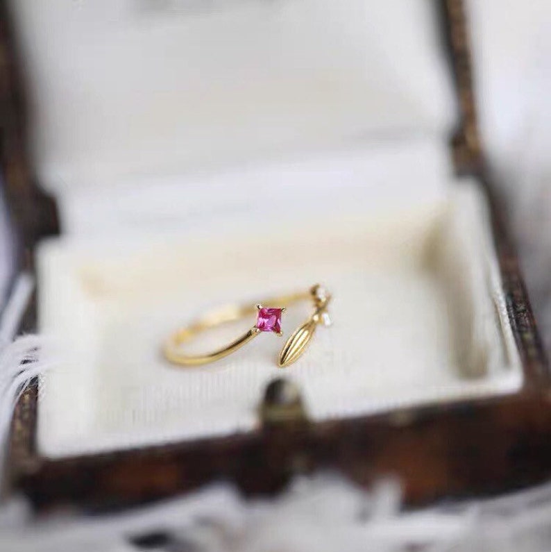 Puede incluir: Un anillo de oro con una piedra cuadrada rosa y un delicado dise&ntilde;o de hoja. El anillo es abierto y ajustable.