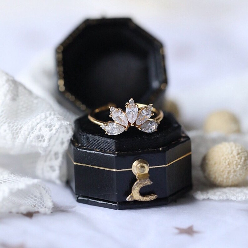 Puede incluir: Un anillo de oro con un dise&ntilde;o delicado que presenta una fila de piedras transparentes en forma de pera dispuestas en forma de corona. El anillo se muestra en una caja de anillo de terciopelo negro.