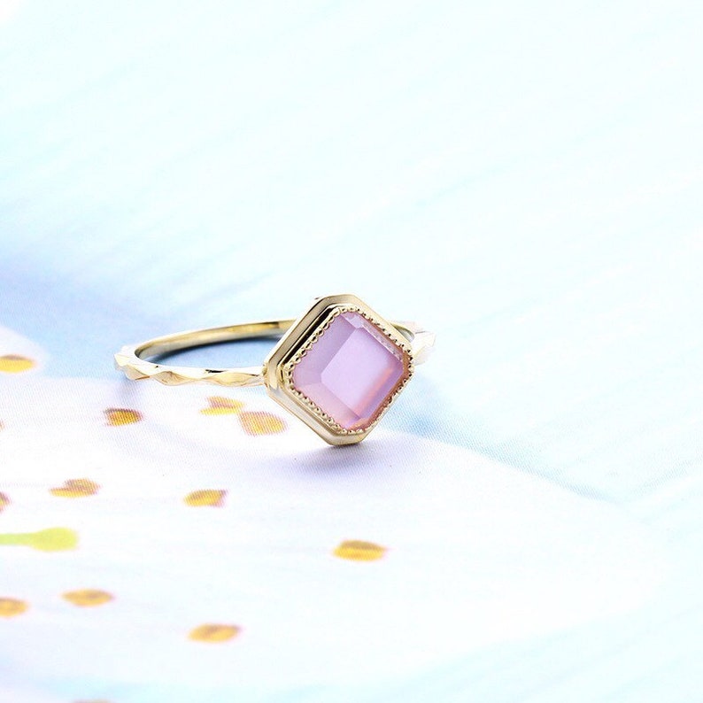 May include: A gold ring with a pink square gemstone set in a decorative gold frame. The ring band is twisted and textured.