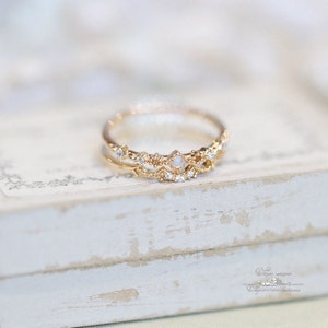 May include: A delicate gold ring with a delicate design featuring small white stones. The ring is set on a white wooden surface.