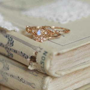 Peut inclure: Une bague dorée composée de deux anneaux reliés, ornée d'une pierre centrale en forme de poire aux reflets irisés et de multiples petites pierres transparentes incrustées dans une structure métallique ajourée et finement travaillée.