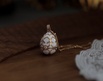 Pode incluir: Um pingente dourado em forma de ovo numa corrente delicada, com acabamento em esmalte branco e padrões intrincados de filigrana dourada, adornado com pequenas pedras transparentes incrustadas por toda a superfície e no topo.