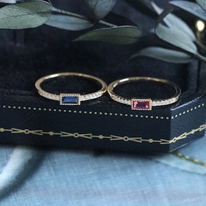 Peut inclure: Deux bagues en or, chacune avec une seule pierre sertie dans la bande. La bague de gauche a une pierre bleue, tandis que la bague de droite a une pierre rouge. Les deux bagues sont ornées d'une rangée de petites pierres claires autour de la bande.