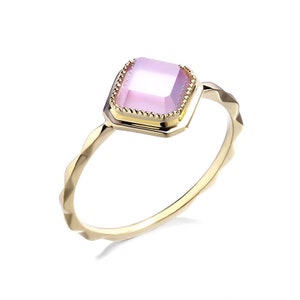 May include: A gold ring with a pink square gemstone set in a decorative frame. The ring band has a textured design.