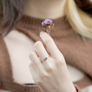 May include: A delicate gold ring featuring a small purple gemstone and smaller clear gemstones on the band.