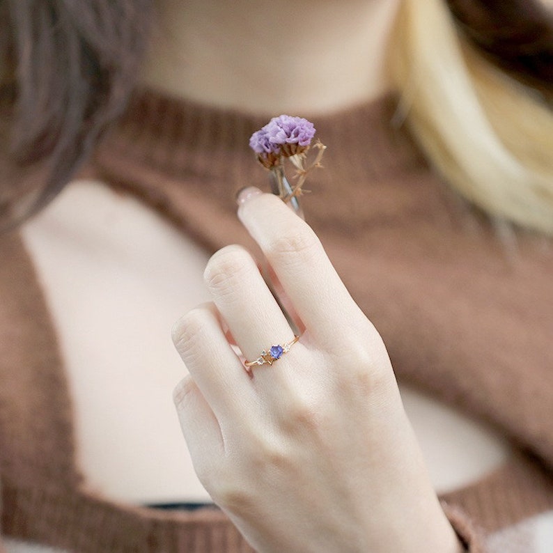 May include: A delicate gold ring featuring a small purple gemstone and smaller clear gemstones on the band.