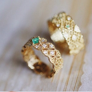 May include: Two gold rings with intricate designs. Both rings feature a small emerald green gemstone set in the center. The rings are on a light wood surface.