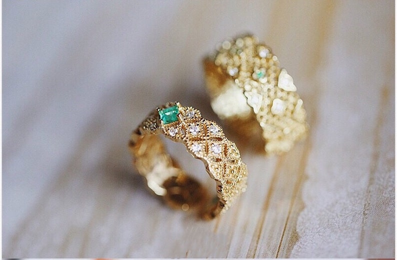 May include: Two gold rings with intricate designs. Both rings feature a small emerald green gemstone set in the center. The rings are on a light wood surface.