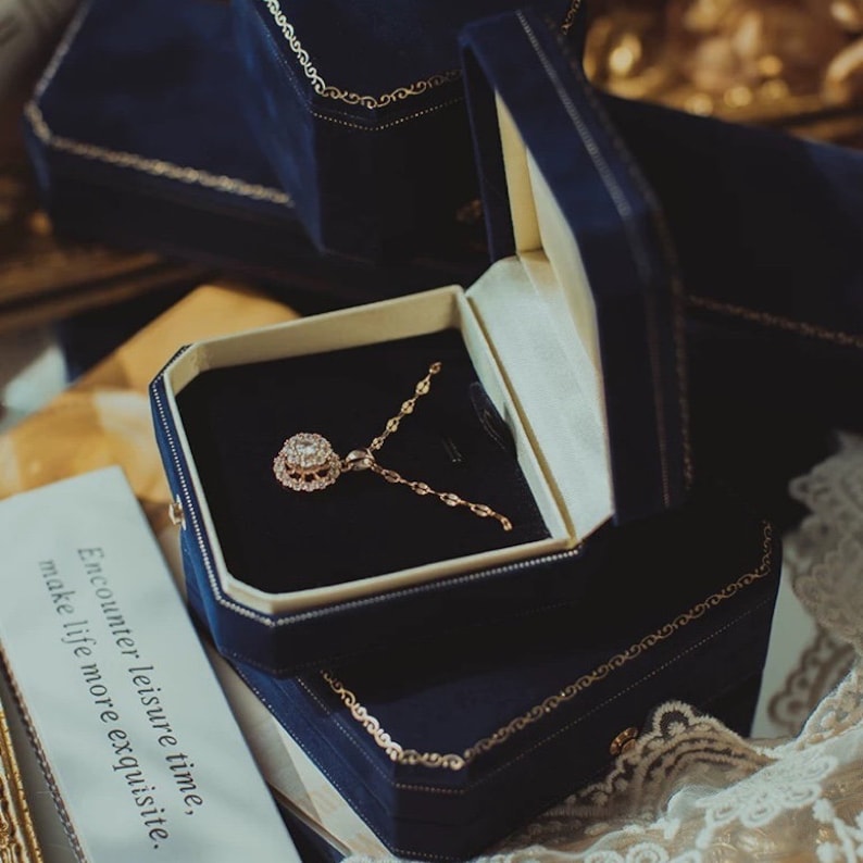 Op de afbeelding: Een gouden ketting met een glinsterende hanger in een blauwe fluwelen juwelendoos. De doos heeft een gouden rand en de woorden "Encounter leisure time, make life more exquisite." staan ​​op de zijkant gedrukt.
