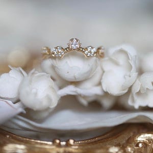 Peut inclure: Une délicate bague en or avec une seule pierre précieuse transparente sertie dans un design en forme de couronne. La bague est ornée de pierres précieuses plus petites le long de la bande, créant un effet scintillant.