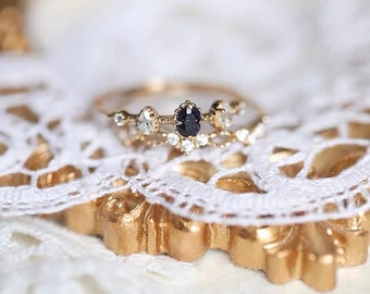 May include: A delicate gold ring with a black oval gemstone surrounded by smaller clear gemstones. The ring is set on a white lace background.
