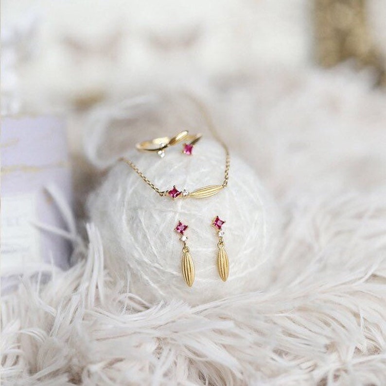 Puede incluir: Un conjunto de joyas de oro que incluye un delicado collar con un dise&ntilde;o de hoja y dos pendientes a juego con peque&ntilde;as piedras rosas. Las joyas se muestran sobre una superficie blanca.