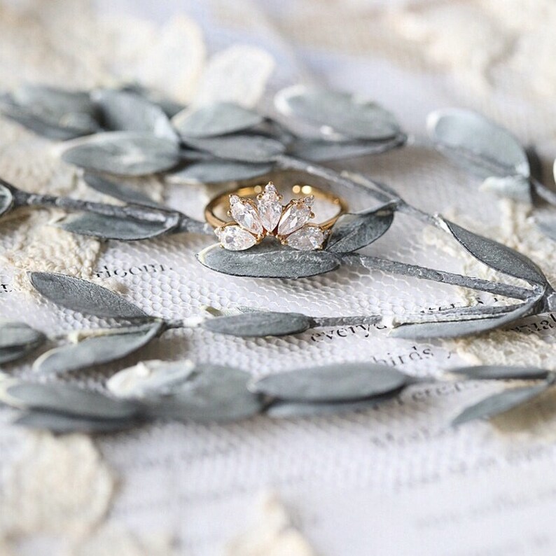 Puede incluir: Un anillo de oro con un dise&ntilde;o delicado que presenta una fila de piedras claras en forma de marquesa. El anillo est&aacute; colocado sobre un fondo de hojas suaves de color verde gris&aacute;ceo.