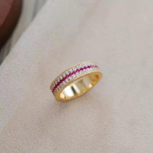 May include: A gold-colored ring with a band of alternating rows of small, round, white and red gemstones. The ring is set against a neutral background, highlighting its design and color.