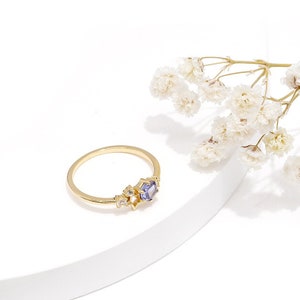 May include: A gold ring featuring a purple gemstone and smaller clear gemstones. The ring is displayed on a white surface.