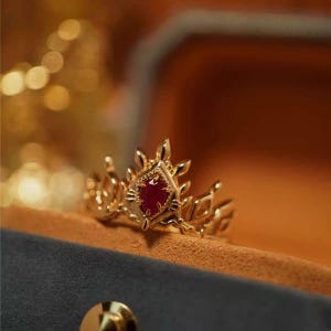 May include: A gold ring featuring a pointed, crown-like design with a central marquise-cut red gemstone. The metal band has intricate, sharp-edged detailing that extends upward, creating a jagged, symmetrical silhouette around the stone.
