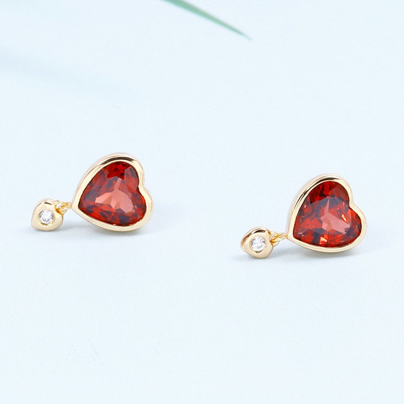 May include: A pair of gold heart-shaped earrings with red gemstone accents and a small clear gemstone dangling from each earring.