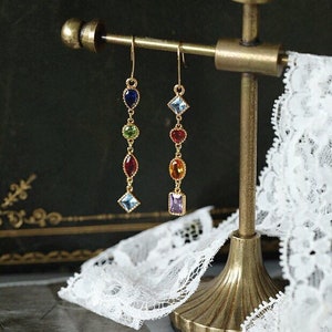 May include: A pair of gold dangle earrings with colourful gemstones. The earrings feature a variety of shapes and sizes of gemstones, including round, square, and oval. The gemstones are arranged in a vertical line on each earring.