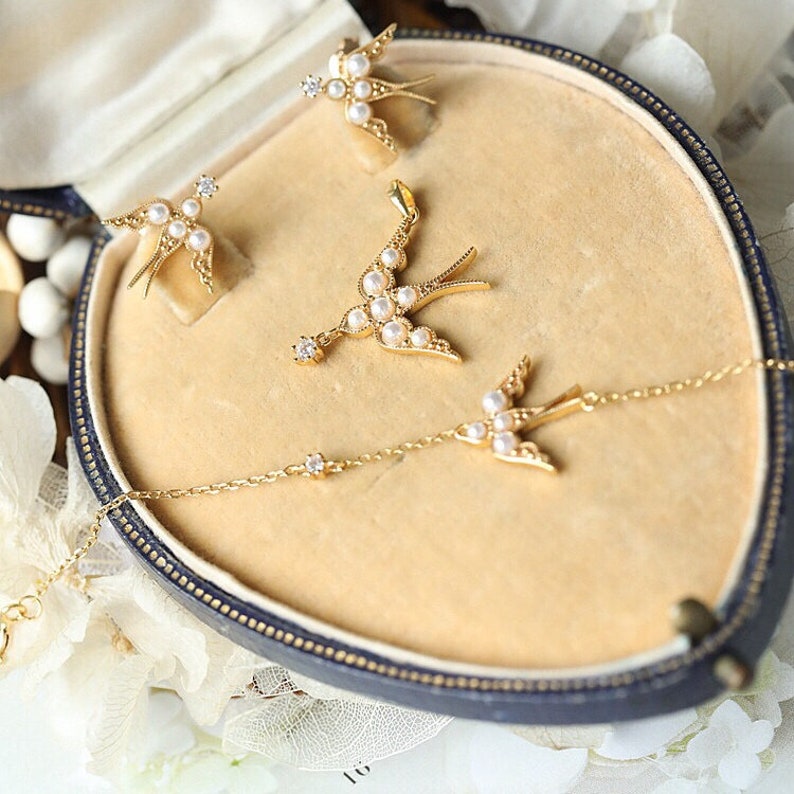 K&ouml;nnte beinhalten: Ein Set aus goldfarbenem Schmuck mit einem zarten Schwalbenmotiv mit Perlenakzenten. Das Set enth&auml;lt eine Halskette, ein Paar Ohrringe und einen Anh&auml;nger.