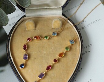May include: A gold necklace with a delicate chain and a pendant featuring a row of colorful gemstones. The pendant is shaped like a teardrop and hangs from a small loop. The necklace is displayed in a velvet-lined jewelry box.