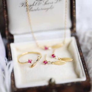 Puede incluir: Un conjunto de delicadas joyas de oro que incluye un anillo, pendientes y un collar. Cada pieza tiene una peque&ntilde;a piedra roja y un dise&ntilde;o en forma de hoja.