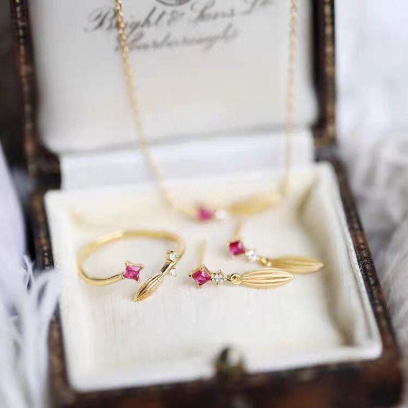 Puede incluir: Un conjunto de delicadas joyas de oro que incluye un anillo, pendientes y un collar. Cada pieza tiene una peque&ntilde;a piedra roja y un dise&ntilde;o en forma de hoja.