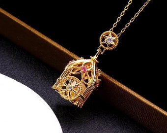 Pode incluir: Um colar de ouro com um pingente detalhado em forma de gaiola de pássaros com pedras preciosas rosa e brancas. O pingente está preso a uma delicada corrente de ouro.