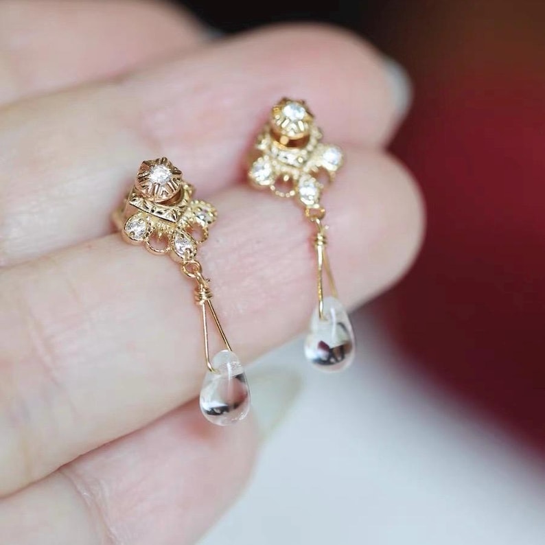 May include: A pair of gold-colored drop earrings. Each earring features a decorative top with small clear stones and a teardrop-shaped clear stone pendant. The earrings are designed with intricate details and a delicate appearance.