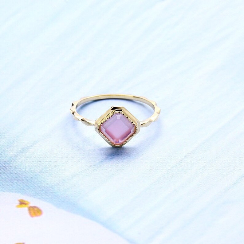 May include: A gold ring with a pink square-shaped gemstone set in a decorative frame.
