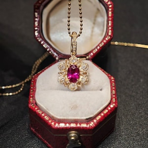 Könnte beinhalten: Eine goldene Halskette mit Anhänger und floralem Design. Der Anhänger hat einen großen, ovalen Rubin-Edelstein, der von Diamanten umgeben ist. Die Halskette wird in einer roten Schmuckschatulle mit weißem Innenraum präsentiert.