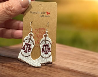 May include: White embroidered cowboy boot earrings with the letters "ATM" in maroon. The earrings are on a brown card that says "Handmade with love".